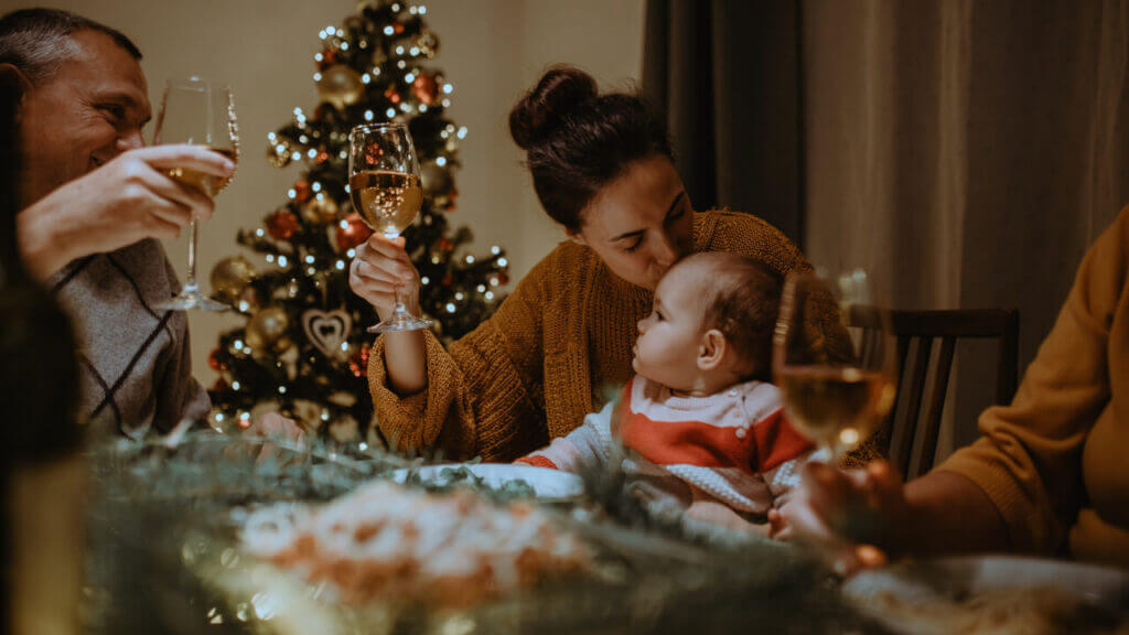 a woman holding a baby raises her glass with the man next to her in front of a Christmas tree stress-free Christmas