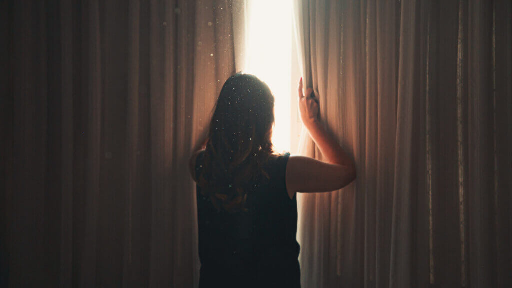 back of woman in a dark room holding curtains slightly parted to look out of the window emotional abuse