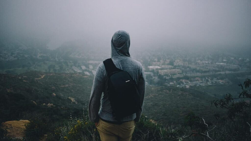 a man in a hoodie with a backpack overlooks a faraway city from a mountain anxious about life