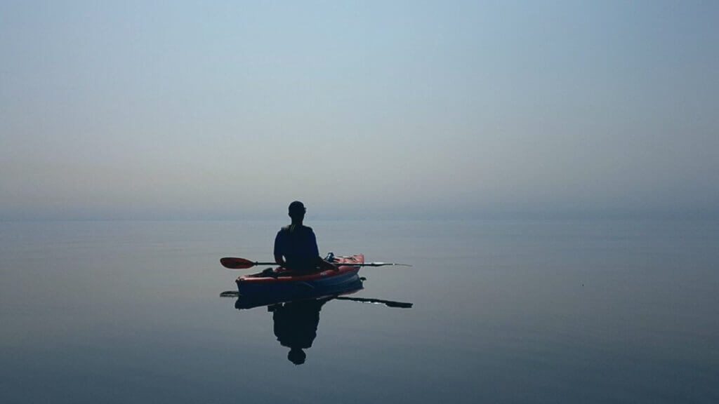 a man in a kayak on a smooth body of water facing the horizon restless mind Catholic Mindfulness