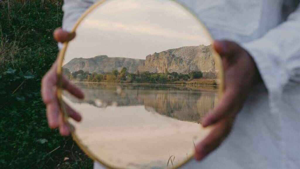 hands hold a small round mirror reflecting a lake and mountain perfectionism